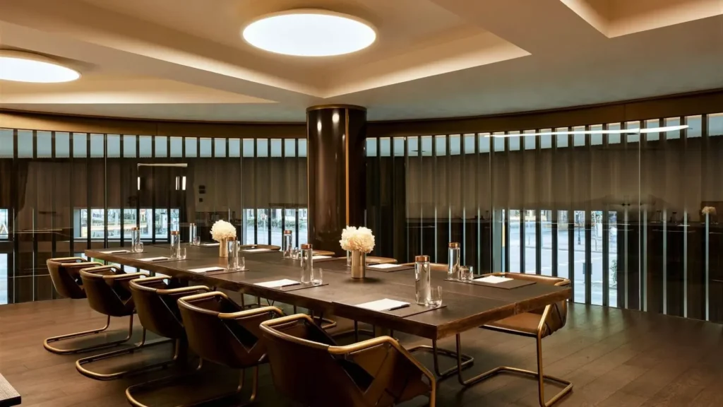 Modern conference room with a long rectangular table, ten chairs, notepads, pens, bottles of water, and vases with white flowers under ceiling lights. Large windows with sheer curtains.